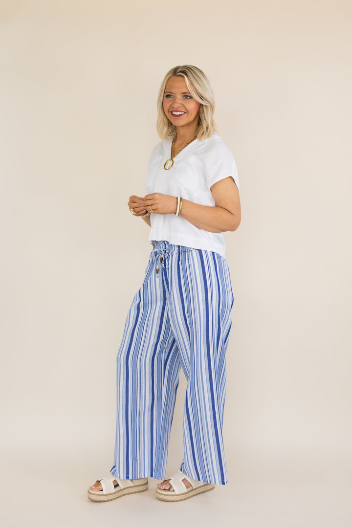 A woman modeling a white short-sleeve top and blue-and-white vertically striped wide-leg pants with beige sandals, standing against a plain beige studio background.