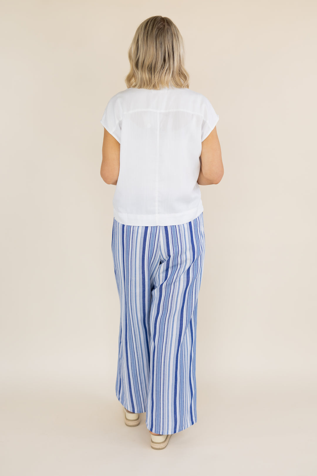 A woman viewed from behind wearing a white short-sleeve top and blue-and-white vertically striped wide-leg pants, standing against a plain beige studio background.