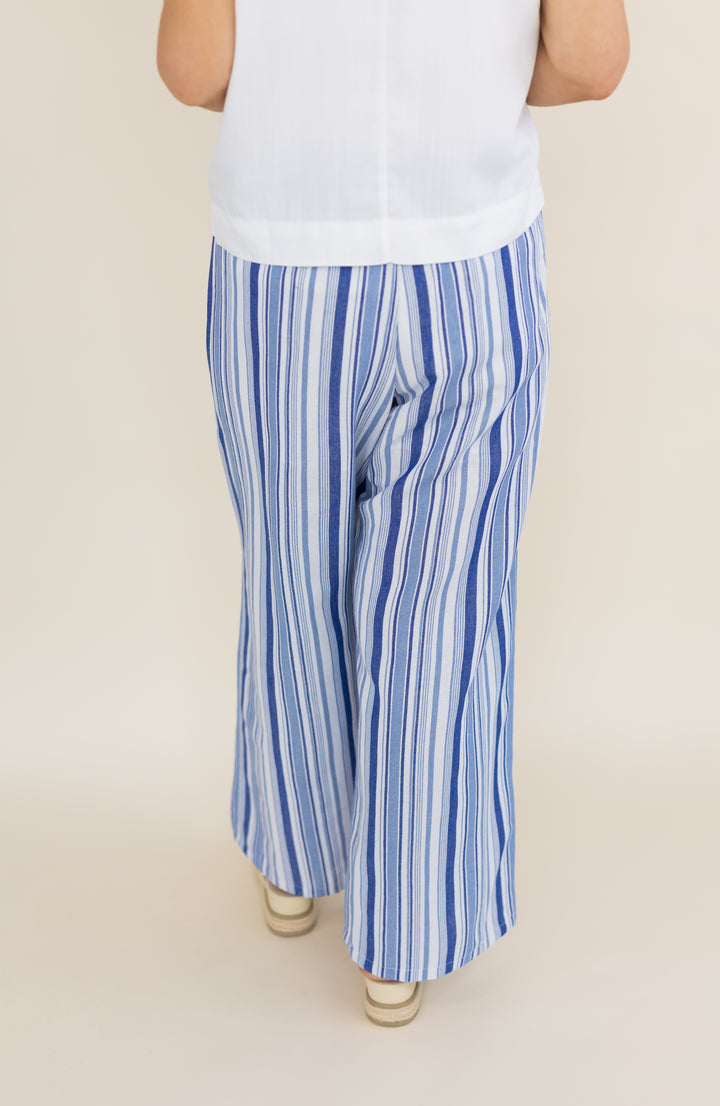 A person shown from the back wearing blue and white vertically striped wide-leg pants, a white sleeveless top, and beige sandals against a plain beige studio background.