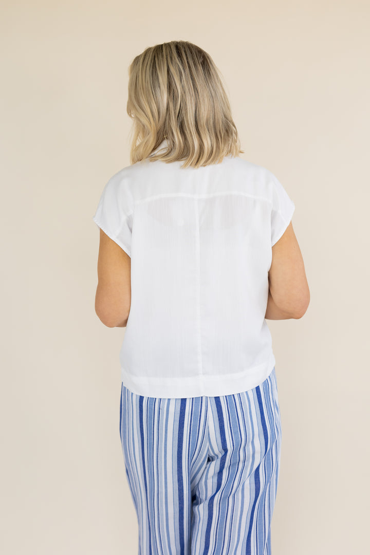 A woman viewed from behind wearing a white short-sleeve blouse and blue-and-white vertically striped pants, standing against a plain beige studio background.