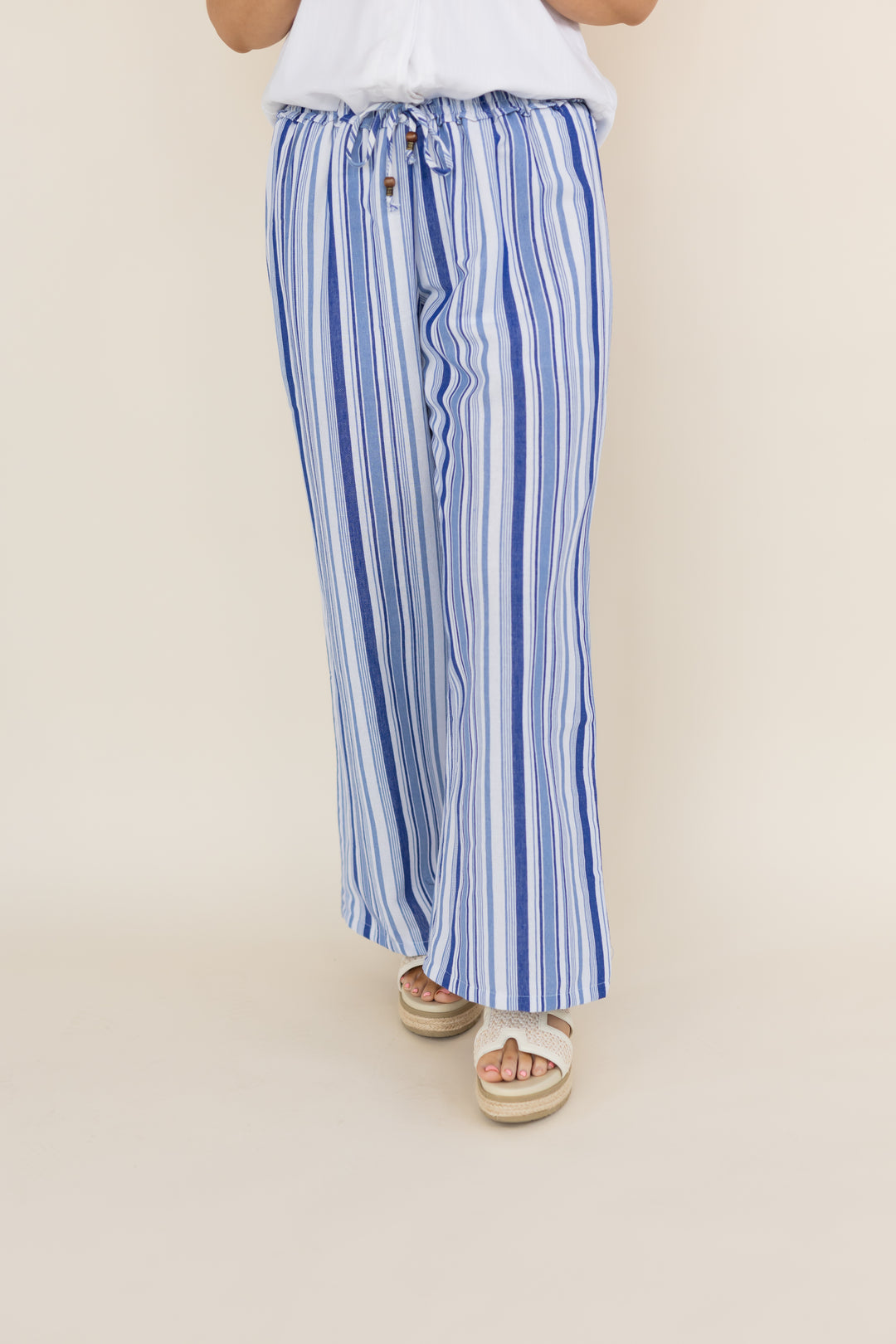 A person wearing blue and white vertically striped wide-leg drawstring pants with a white top and white espadrille sandals, shown from the waist down against a plain beige studio background.