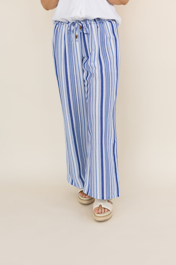 A person wearing blue and white vertically striped wide-leg drawstring pants with a white top and white espadrille sandals, shown from the waist down against a plain beige studio background.