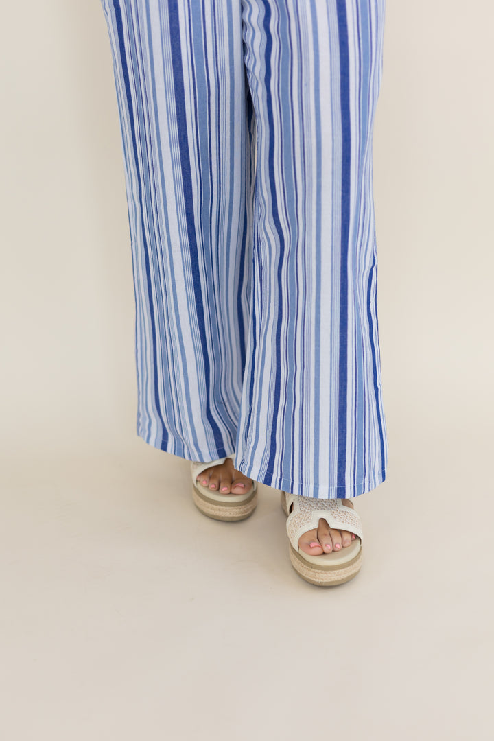 A person wearing blue and white vertically striped wide-leg pants and white espadrille sandals, shown from the knees down against a plain light beige studio background.