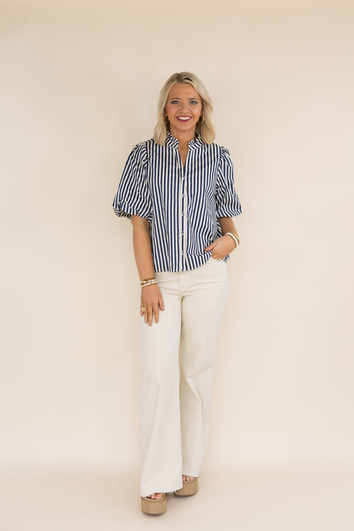 A woman modeling a short-sleeve button-up shirt with vertical navy and white stripes, paired with white wide-leg pants and beige platform sandals, standing against a solid cream background.