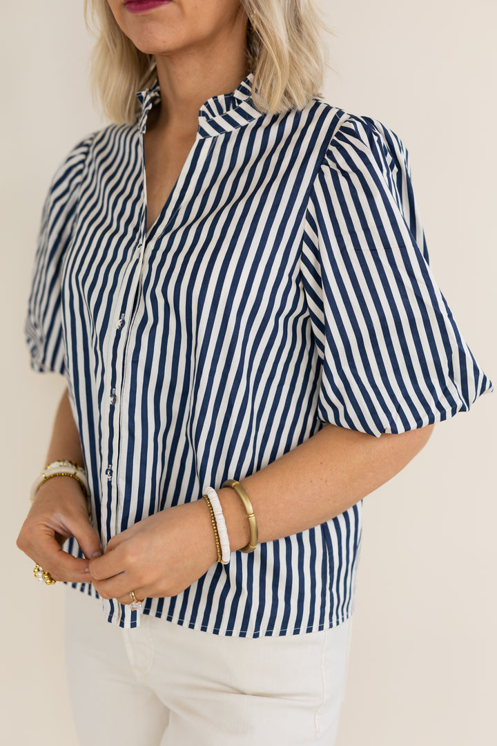 A woman wearing a navy and white vertically striped short-sleeve blouse with puff sleeves, paired with white pants, against a plain light beige background.