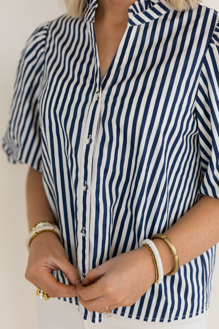 A close-up of a woman wearing a short-sleeve button-front shirt with vertical navy and white stripes, adjusting the front buttons. She is wearing white and gold bracelets on both wrists.