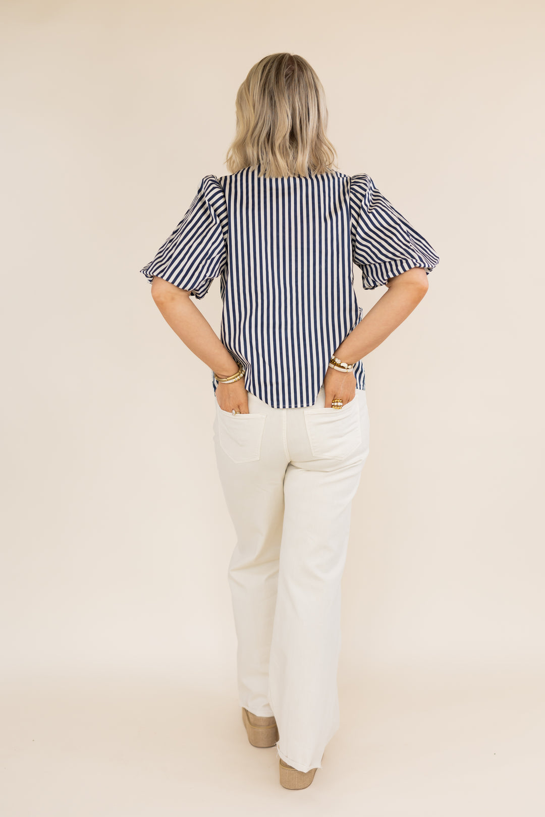 A woman shown from the back wearing a navy and white vertically striped puff-sleeve blouse with white pants, standing against a solid beige studio background.
