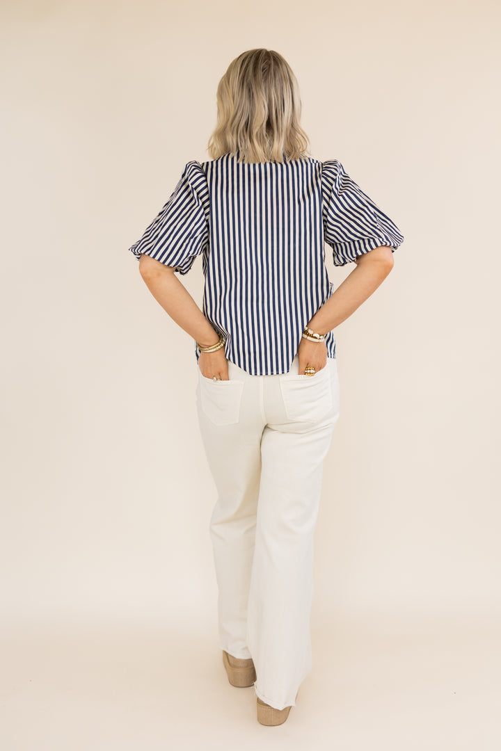 A woman shown from the back wearing a navy and white vertically striped puff-sleeve blouse with white pants, standing against a solid beige studio background.