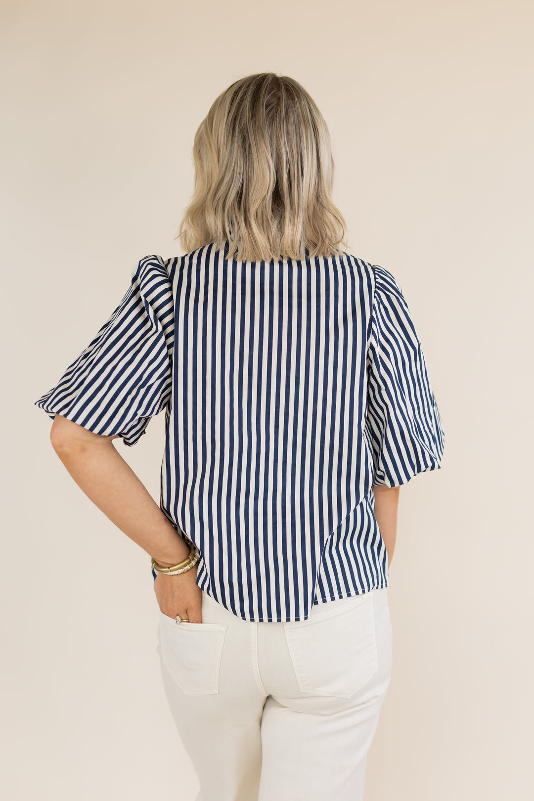 A woman viewed from behind wearing a navy and white vertically striped blouse with ruffled short sleeves and white pants, standing against a plain beige background.