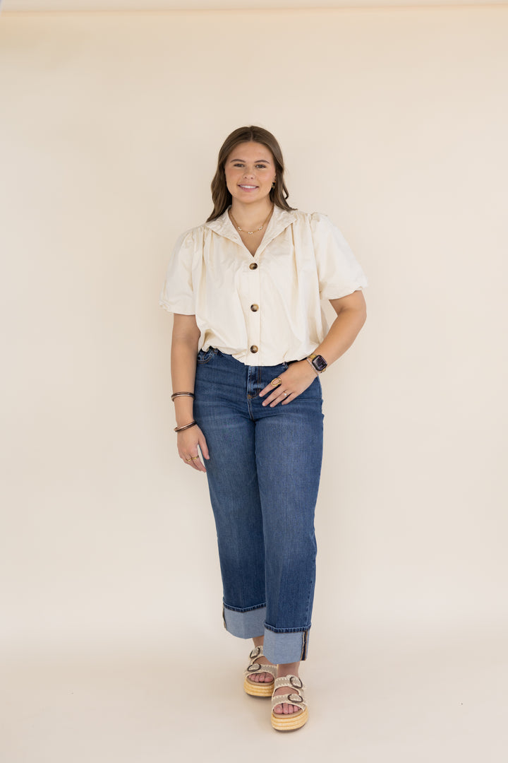 A woman modeling a cream short-sleeve button-up shirt and blue cuffed jeans, standing against a plain beige studio background.