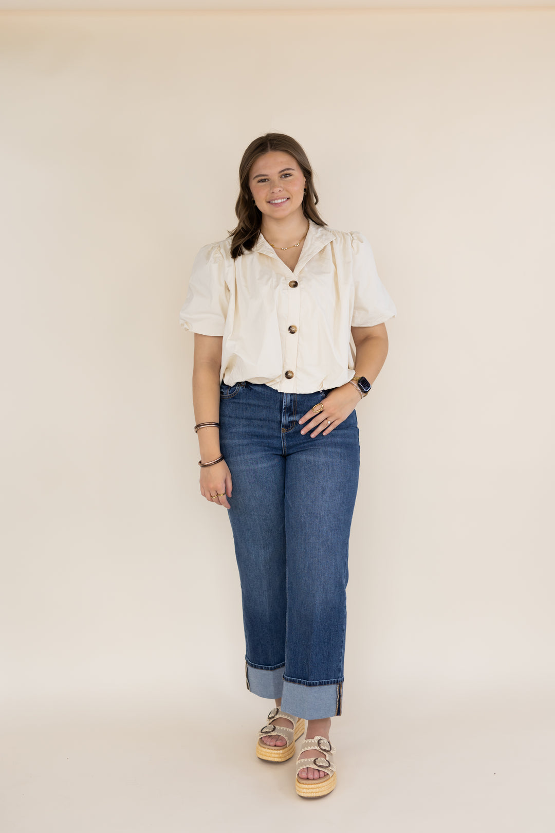 A young woman stands against a plain beige studio background wearing a cream-colored short-sleeve button-up shirt, blue cropped jeans, and pinkish-brown platform sandals.