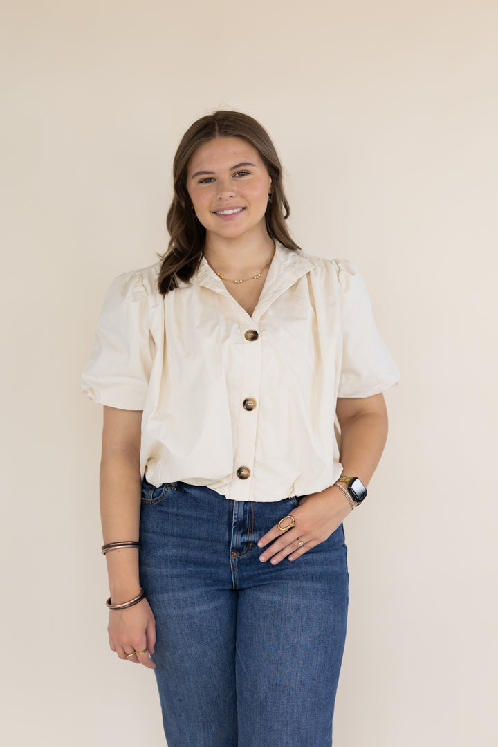 A woman modeling a cream-colored short-sleeve blouse with puffed shoulders and brown buttons, paired with blue jeans, against a solid beige background.