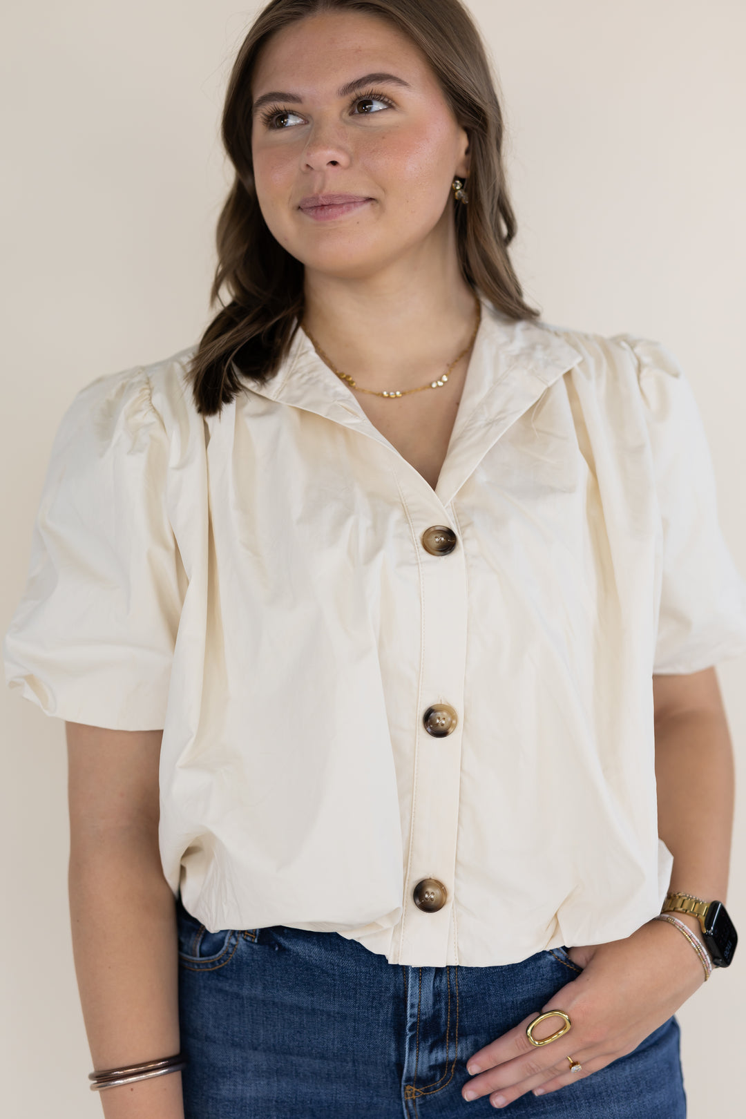 A woman wearing a cream-colored short-sleeve blouse with puffed sleeves and brown buttons, paired with blue jeans, against a plain beige background.