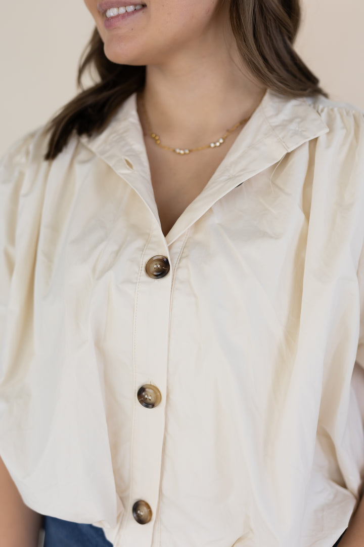 A close-up of a woman wearing a cream-colored short-sleeve button-up blouse with brown buttons and a delicate gold necklace.