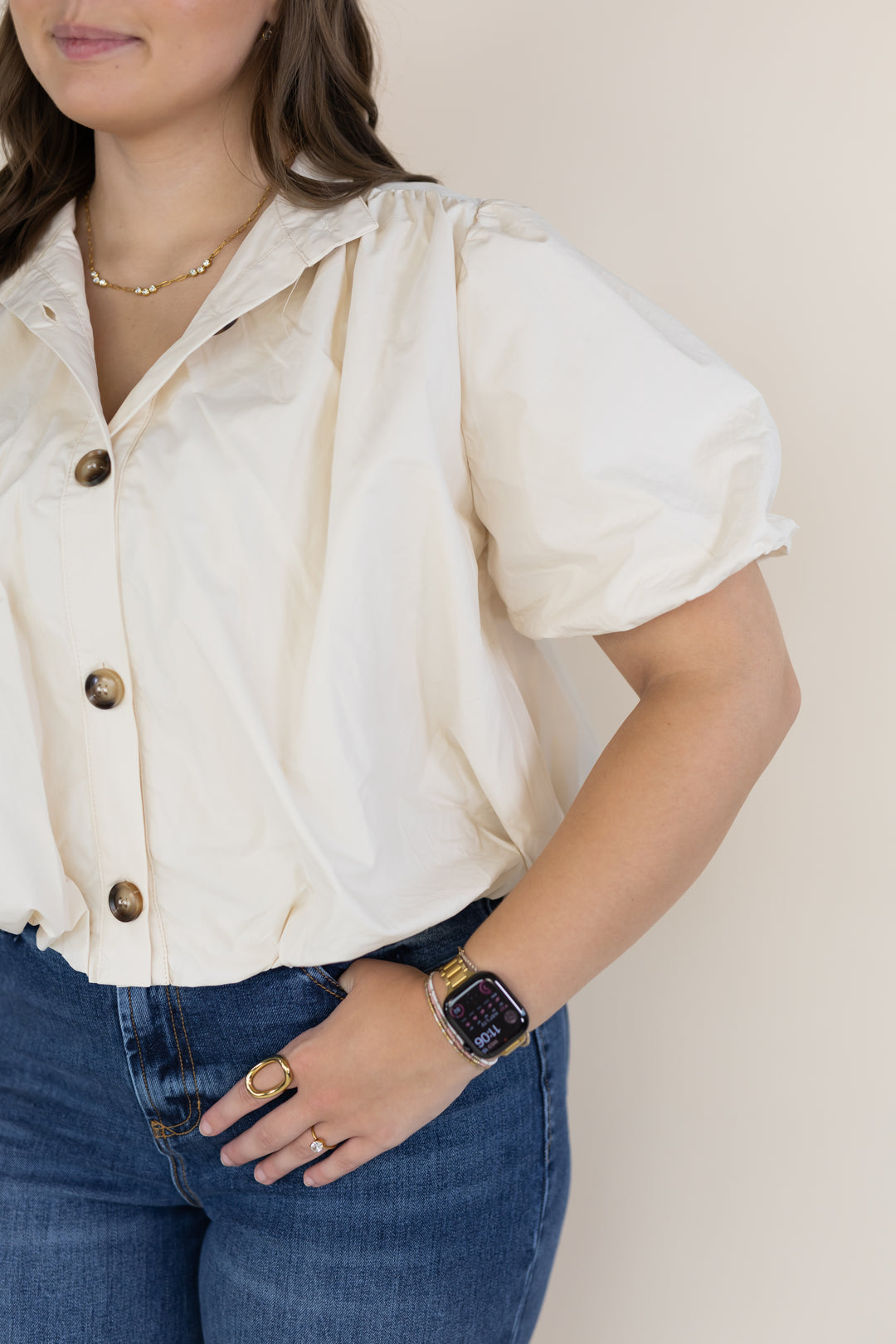 A woman wearing a cream puff-sleeve button-up blouse with dark brown buttons, paired with blue jeans, shown from the chest down against a plain beige background.