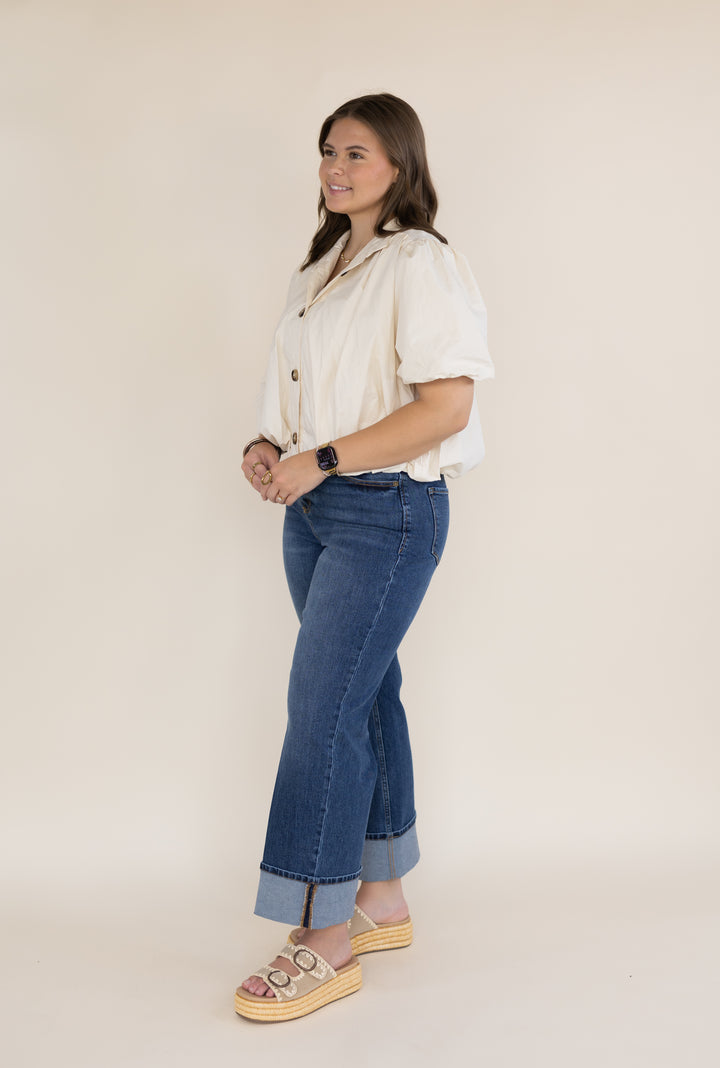 A woman modeling a cream short-sleeve button-up shirt and blue cuffed jeans with platform sandals, standing against a plain beige studio background.