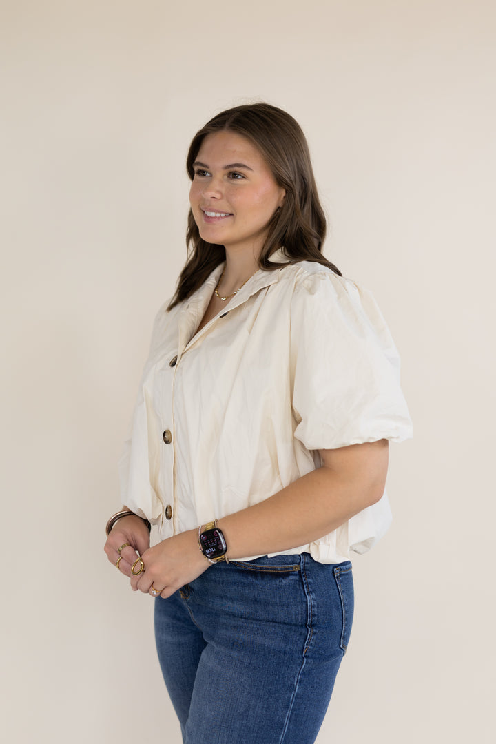 A woman modeling a cream-colored short-sleeve button-up shirt with puffed sleeves, paired with blue jeans, against a plain beige studio background.