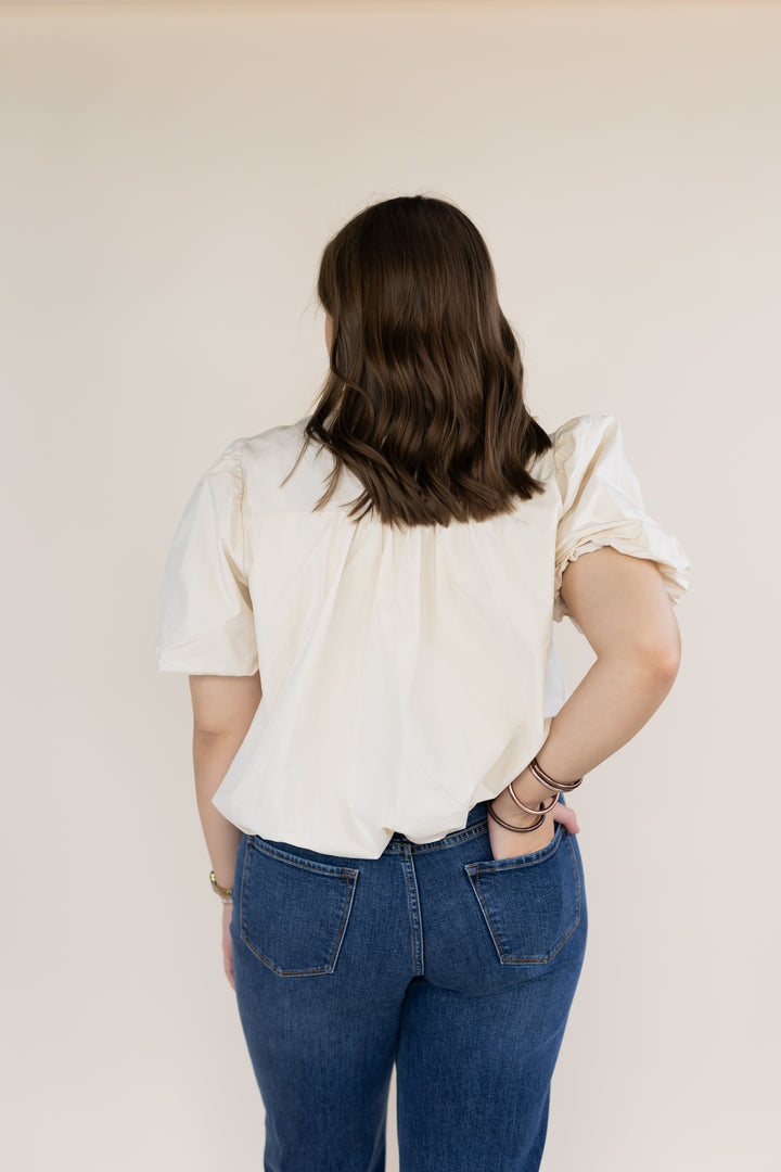 A woman viewed from behind wearing a cream-colored short-sleeve blouse with a gathered back and blue jeans, standing against a plain beige studio background.