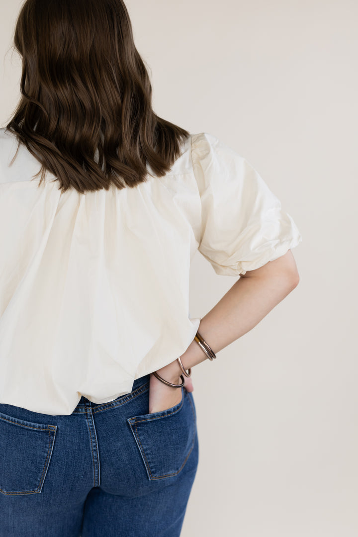 A woman shown from behind wearing a cream puff-sleeve blouse with bubble hem paired with blue jeans, with her hand in her pocket, against a plain light beige background.