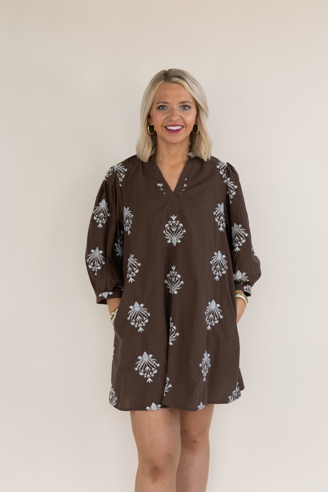A woman with blonde hair wearing a brown long-sleeve tunic dress with a white floral block print pattern, standing against a plain beige background.