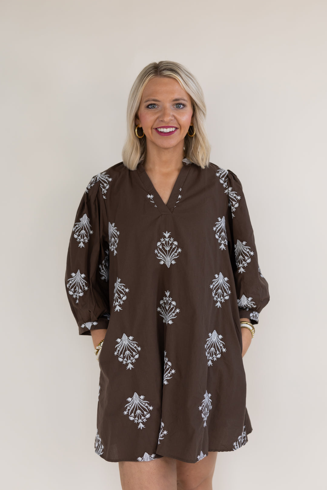 A smiling blonde woman wearing a brown long-sleeve tunic dress with white floral block prints, standing against a plain light beige background.