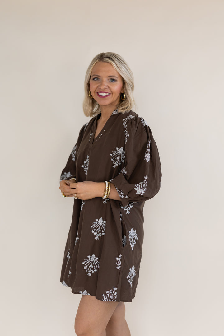 A smiling blonde woman wearing a brown long-sleeve mini dress with a white floral block print, standing against a plain beige studio background.