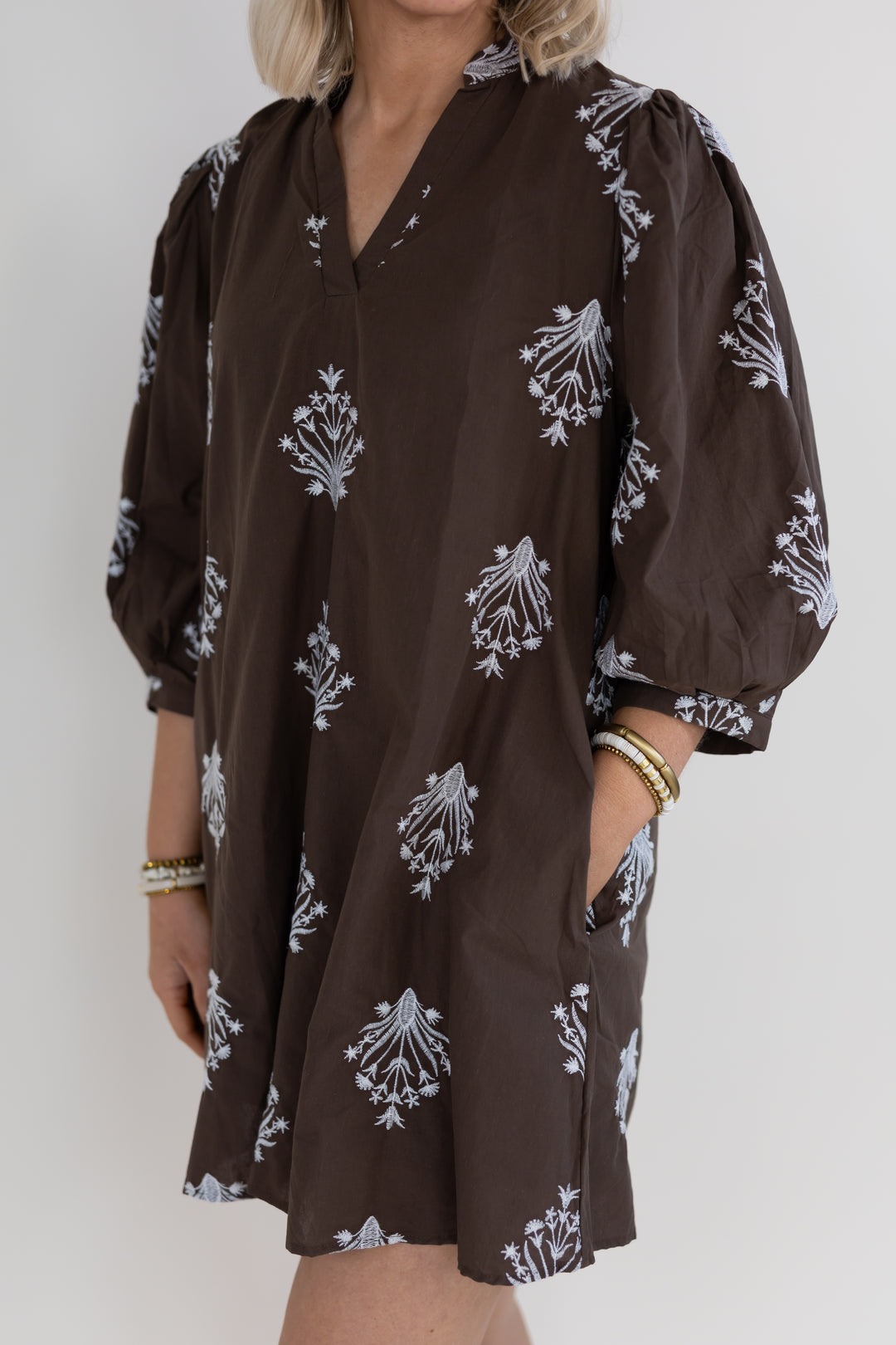 A woman wearing a brown long sleeve mini dress with a white floral pattern, featuring puff sleeves and side pockets, photographed against a plain light background.