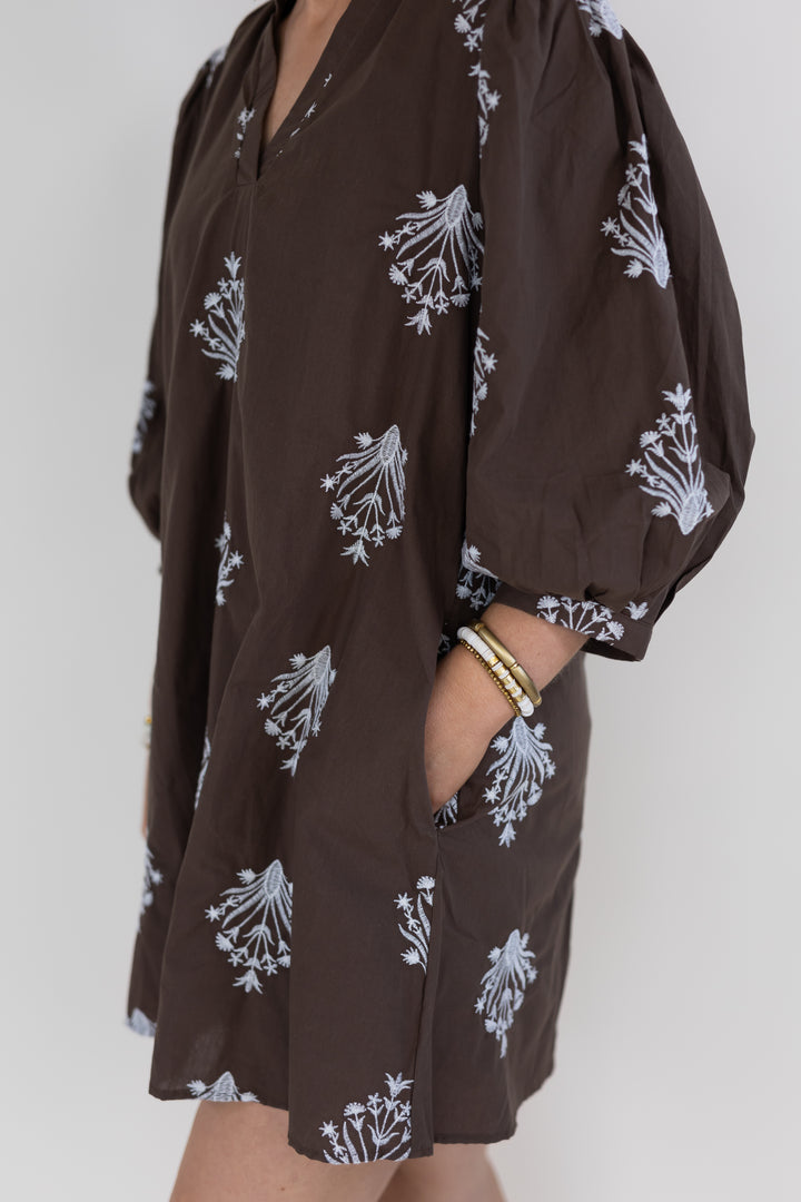 A close-up of a person wearing a brown short-sleeve tunic dress with a white floral print, featuring puff sleeves and side pockets.