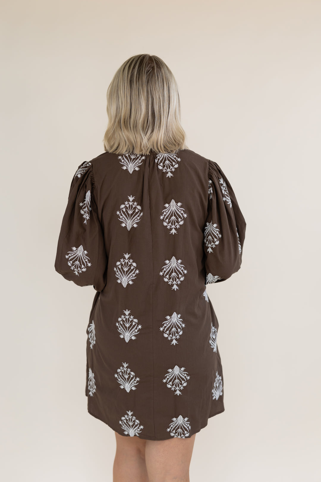 A woman viewed from behind wearing a brown puff-sleeve dress with a white damask pattern, set against a plain beige background.