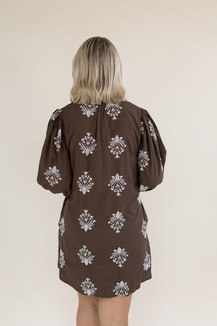 A woman viewed from behind wearing a brown puff-sleeve dress with a white damask pattern, set against a plain beige background.