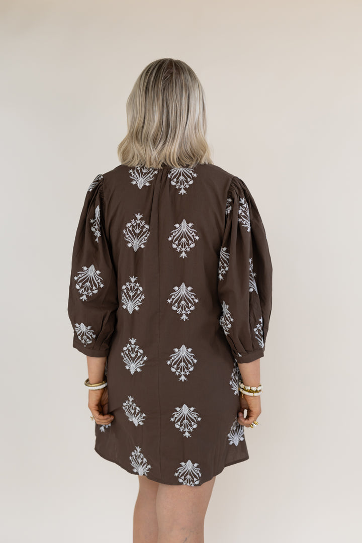 A woman viewed from behind wearing a brown puff-sleeve dress with a white floral pattern, set against a plain light beige background.