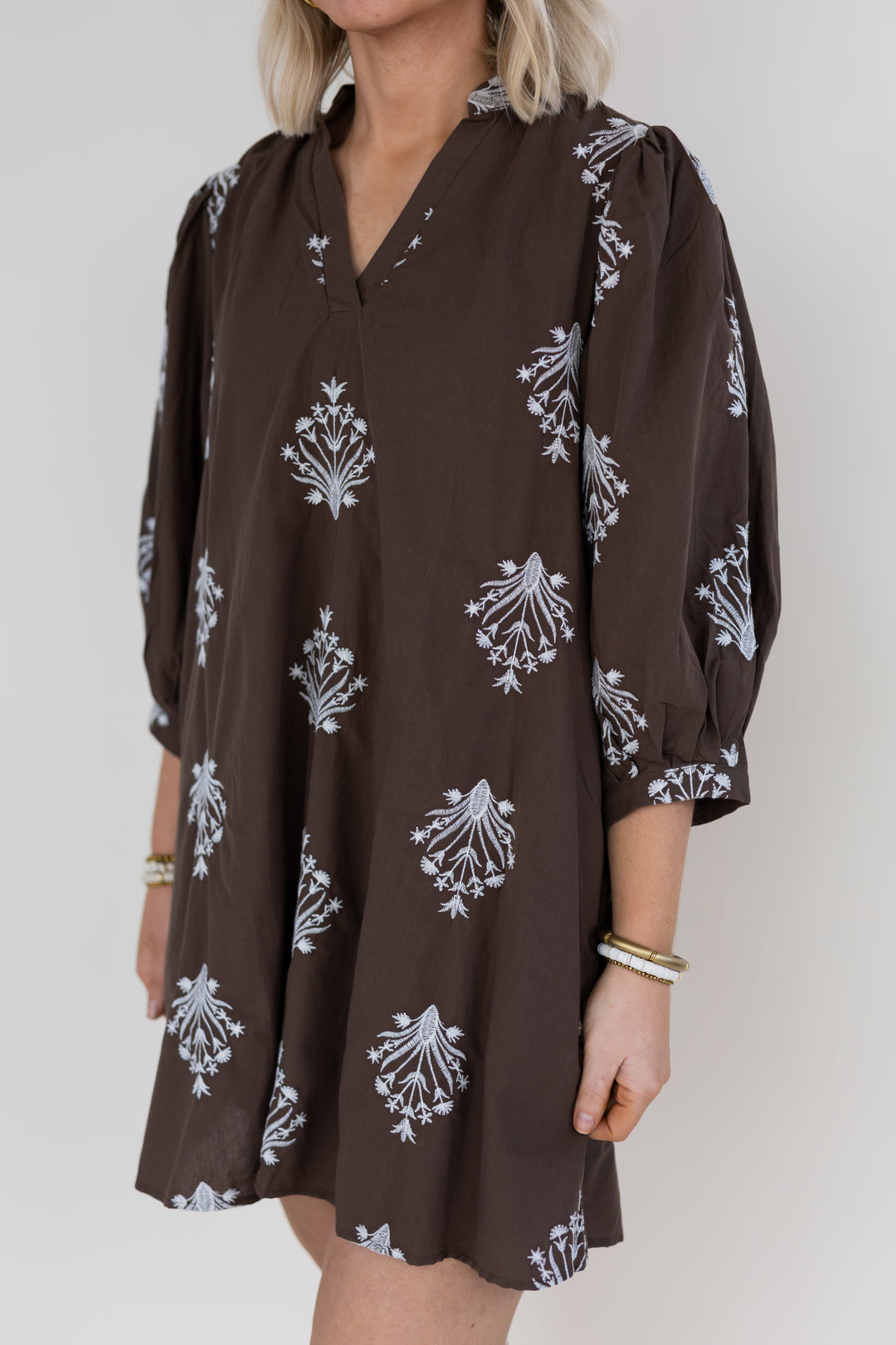 A woman wearing a dark brown tunic dress with a white floral block print pattern, featuring puff sleeves and a V-neckline, shown against a plain light gray background.