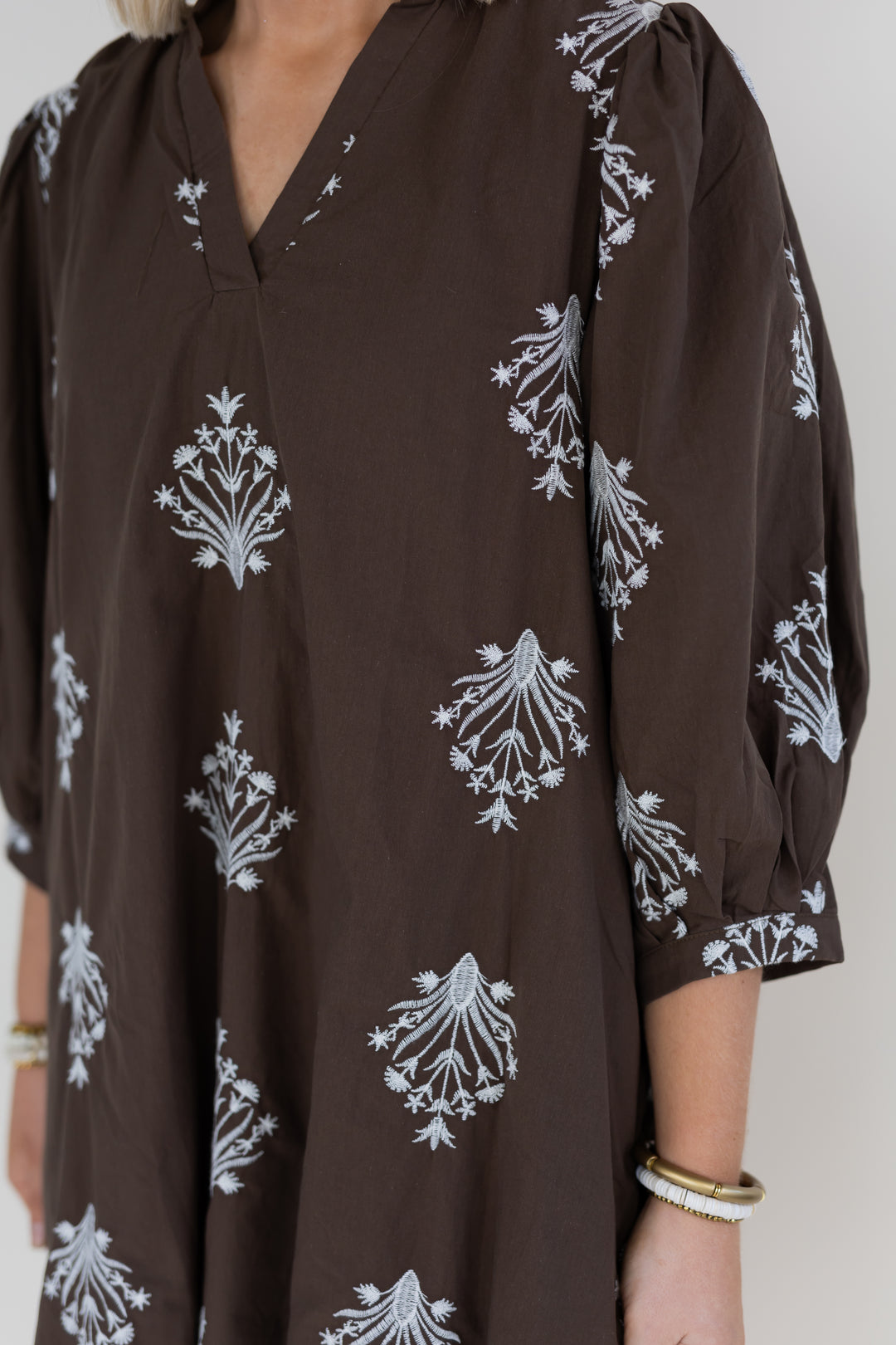 A close-up of a woman wearing a dark brown tunic dress with white floral block prints, featuring puff sleeves and a V-neckline.