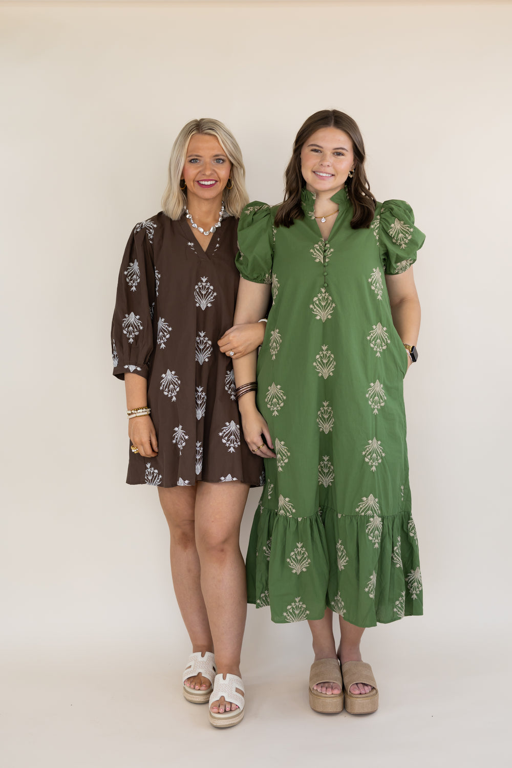 Two women modeling bohemian-style dresses against a plain cream background. The woman on the left wears a brown short dress with white floral prints, and the woman on the right wears an olive green midi dress with white floral prints and puff sleeves.