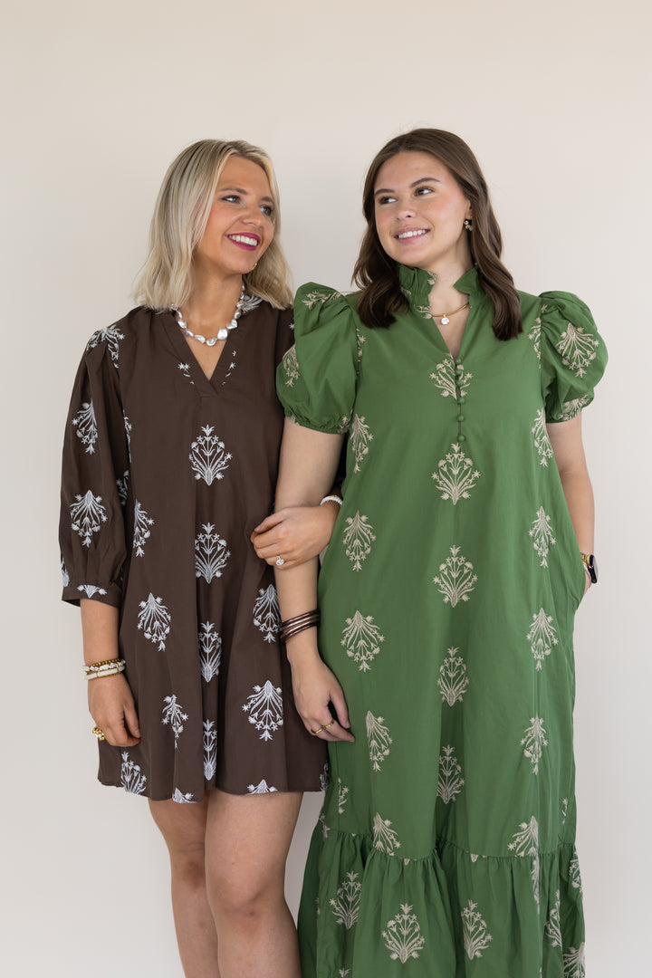 Two women modeling patterned dresses against a plain light beige background. The woman on the left wears a brown short dress with white floral motifs, and the woman on the right wears an olive green long dress with a similar white floral print.