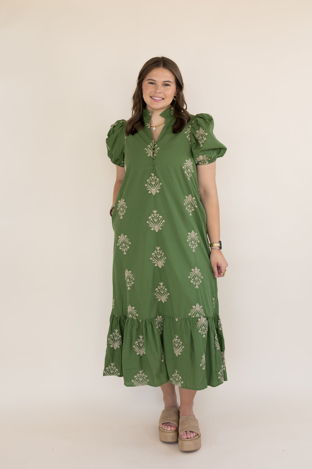 A woman modeling a green midi dress with puff sleeves, a keyhole neckline, and beige floral embroidery, standing against a plain cream background.
