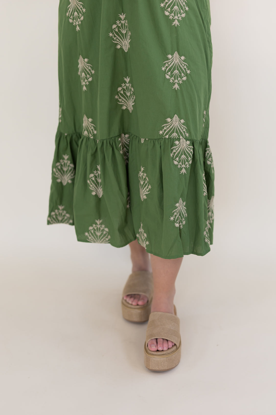 Close-up of green embroidered midi dress with ivory floral detailing, ruffle hem, and neutral platform sandals