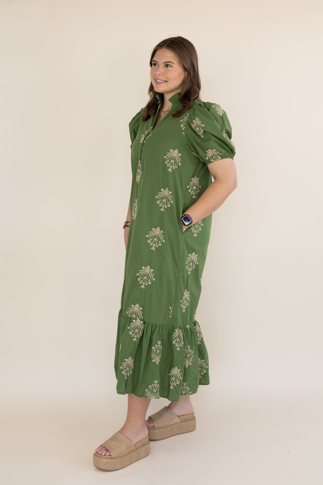 A woman modeling a olive green midi dress with a beige floral pattern, puff sleeves, and a ruffled hem, standing against a plain beige studio background.