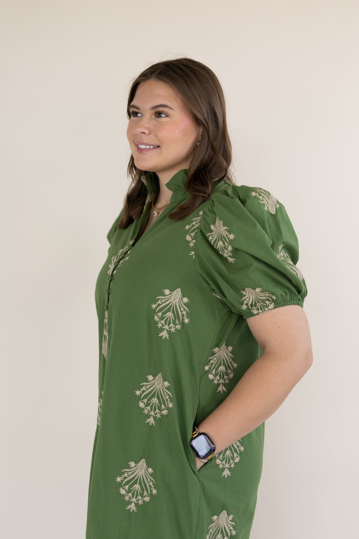 A woman wearing a green short-sleeve button-up dress with beige floral block prints, standing against a plain light beige background.