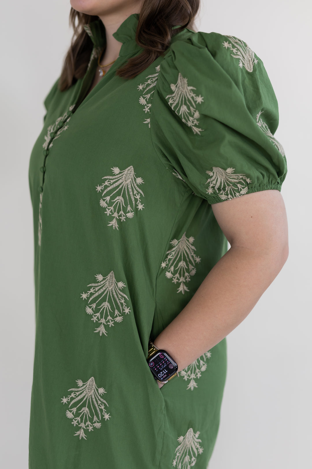 A woman wearing an olive green puff-sleeve maxi dress with beige floral block prints, shown from the side against a plain light gray background.