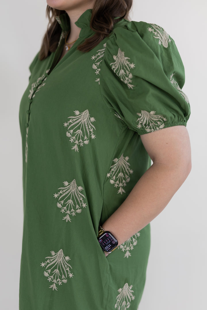 A woman wearing an olive green puff-sleeve maxi dress with beige floral block prints, shown from the side against a plain light gray background.
