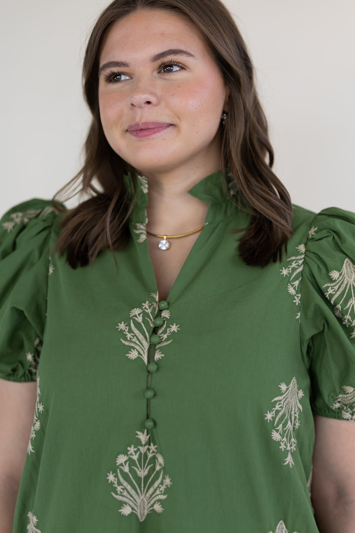 A woman wearing a green short sleeve maxi dress with white floral embroidery and puff sleeves, photographed against a plain light background.
