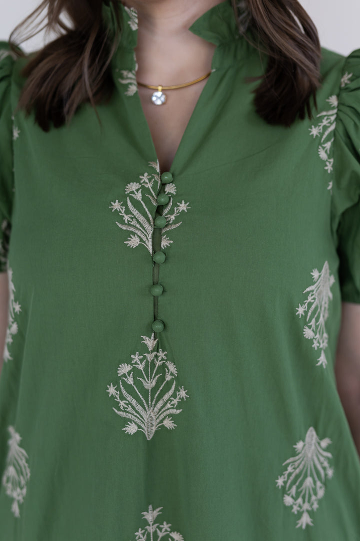 A close-up of a woman wearing an olive short-sleeve maxi dress with white floral embroidery and a buttoned V-neckline.
