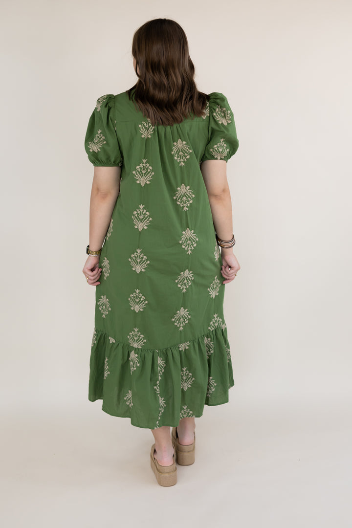 A woman viewed from the back wearing a green midi dress with puff sleeves, a gathered neckline, and an allover beige floral pattern. She is standing against a plain, light beige studio background.