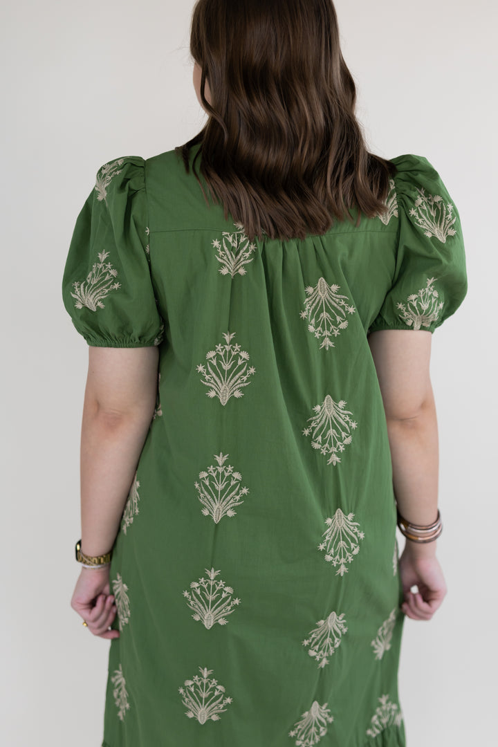 A woman viewed from behind wearing a green dress with beige floral block prints and puff sleeves, set against a plain light gray background.