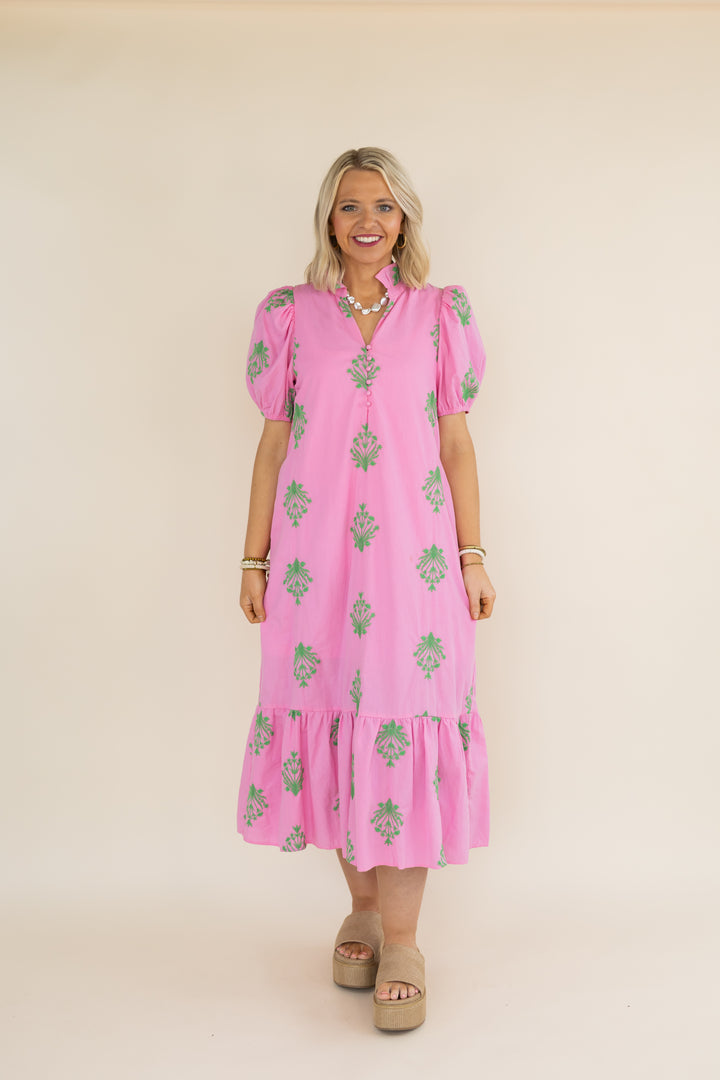 A woman modeling a pink midi dress with green floral prints, puff sleeves, and a tiered hem, standing against a solid beige studio background.