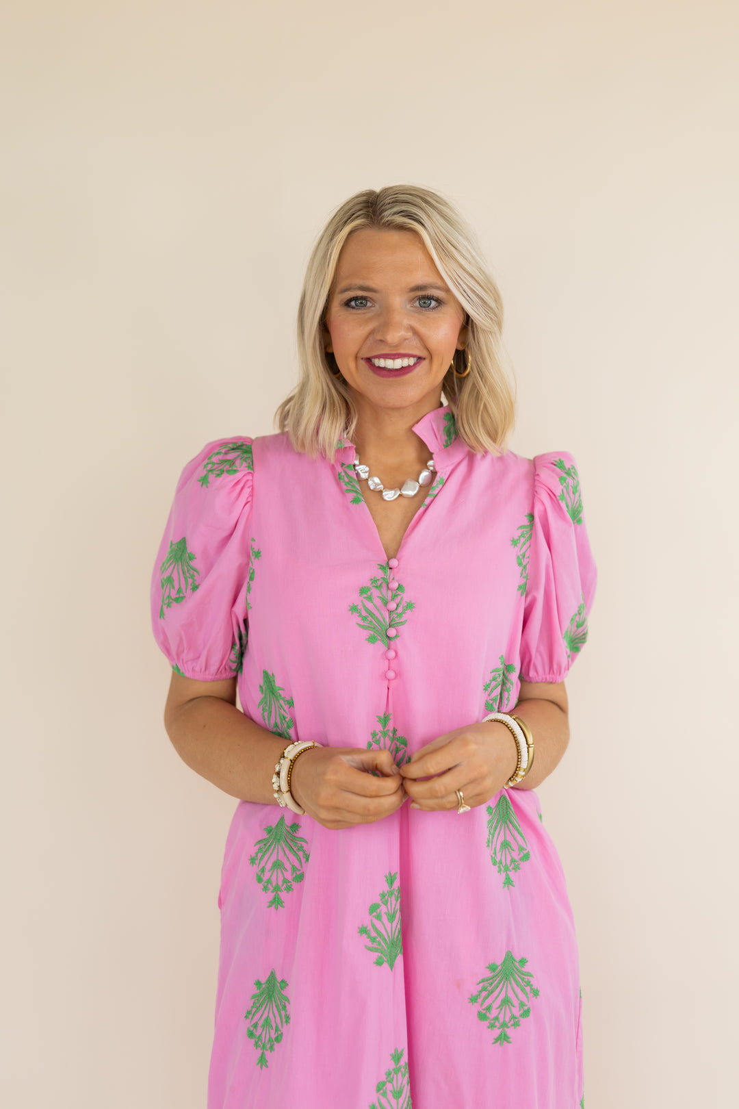 A smiling blonde woman wearing a pink puff-sleeve dress with green floral block prints, standing against a plain beige background.