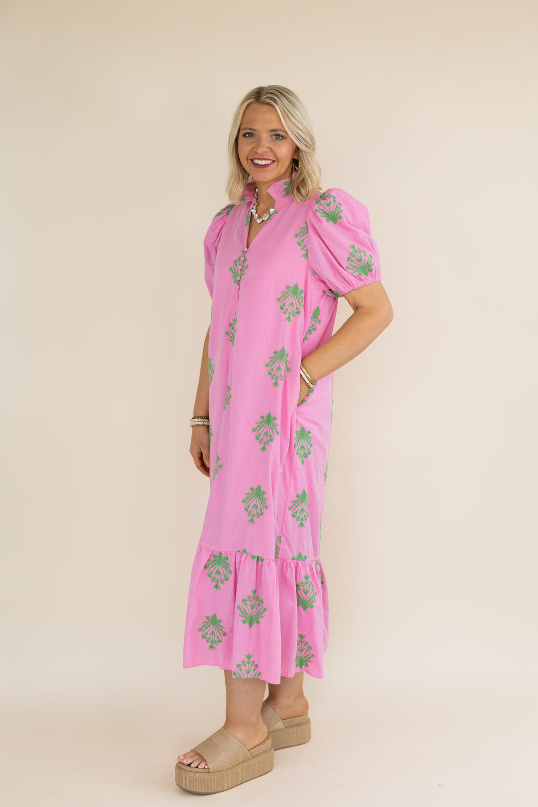 A woman modeling a pink midi dress with green floral prints, puff sleeves, and a tiered hem, standing against a solid beige studio background.