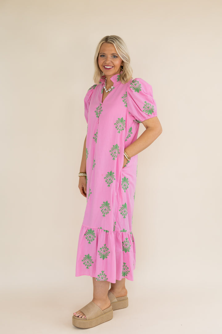 A woman modeling a pink midi dress with green floral prints, puff sleeves, and a tiered hem, standing against a solid beige studio background.