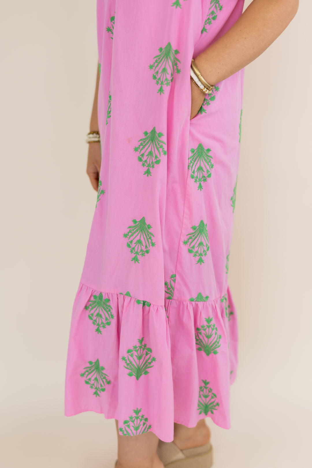 A close-up of a pink short sleeve  dress with green floral block-print patterns and a ruffled hem, shown on a model against a light beige background.